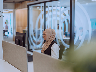 Image showing In a modern office, a young Muslim entrepreneur wearing a hijab sits confidently and diligently works on her computer, embodying determination, creativity, and empowerment in the business world