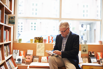 Image showing Senior man, library and reading book for literature, knowledge or story at bookstore. Mature male person, author or bookworm sitting by bookshelf for novel, learning or wisdom in shopping or leisure