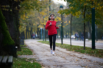 Image showing Young beautiful woman running in autumn park and listening to music with headphones on smartphone