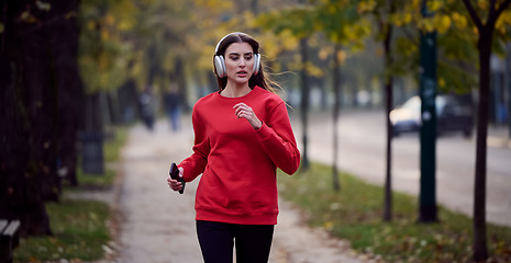 Image showing Young beautiful woman running in autumn park and listening to music with headphones on smartphone