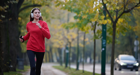 Image showing Young beautiful woman running in autumn park and listening to music with headphones on smartphone