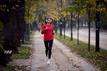 Image showing Young beautiful woman running in autumn park and listening to music with headphones on smartphone