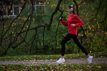 Image showing Young beautiful woman running in autumn park and listening to music with headphones on smartphone