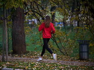 Image showing Young beautiful woman running in autumn park and listening to music with headphones on smartphone