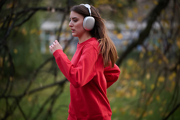 Image showing Young beautiful woman running in autumn park and listening to music with headphones on smartphone