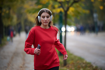 Image showing Young beautiful woman running in autumn park and listening to music with headphones on smartphone