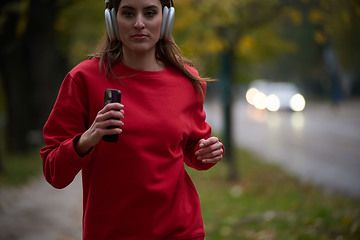 Image showing Young beautiful woman running in autumn park and listening to music with headphones on smartphone