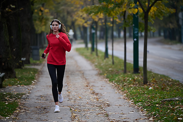 Image showing Young beautiful woman running in autumn park and listening to music with headphones on smartphone