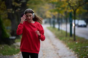 Image showing Young beautiful woman running in autumn park and listening to music with headphones on smartphone