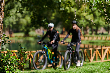 Image showing A blissful couple, adorned in professional cycling gear, enjoys a romantic bicycle ride through a park, surrounded by modern natural attractions, radiating love and happiness