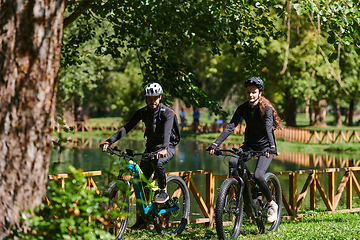 Image showing A blissful couple, adorned in professional cycling gear, enjoys a romantic bicycle ride through a park, surrounded by modern natural attractions, radiating love and happiness