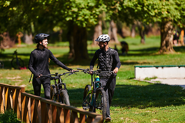 Image showing A blissful couple, adorned in professional cycling gear, enjoys a romantic bicycle ride through a park, surrounded by modern natural attractions, radiating love and happiness