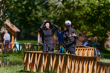 Image showing A blissful couple, adorned in professional cycling gear, enjoys a romantic bicycle ride through a park, surrounded by modern natural attractions, radiating love and happiness