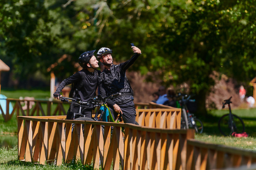 Image showing A blissful couple, adorned in professional cycling gear, enjoys a romantic bicycle ride through a park, surrounded by modern natural attractions, radiating love and happiness