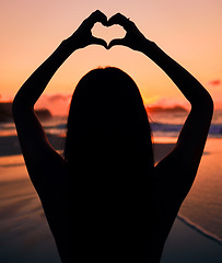 Image showing Woman, silhouette and sunset by sea with heart hands for love, care and kindness with sign, summer and dusk. Girl, person and ocean with symbol, emoji or icon on vacation, beach and outdoor in dark