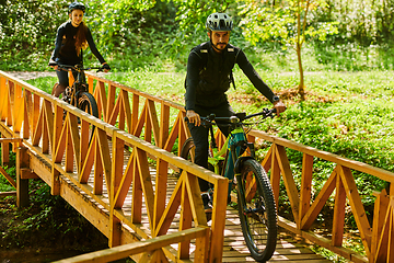 Image showing A blissful couple, adorned in professional cycling gear, enjoys a romantic bicycle ride through a park, surrounded by modern natural attractions, radiating love and happiness