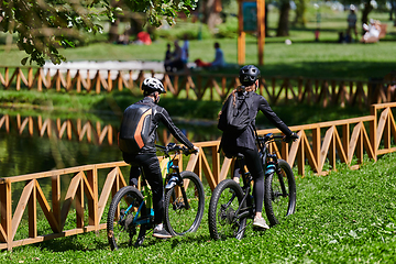 Image showing A blissful couple, adorned in professional cycling gear, enjoys a romantic bicycle ride through a park, surrounded by modern natural attractions, radiating love and happiness