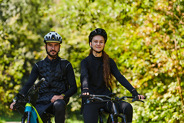 Image showing A blissful couple, adorned in professional cycling gear, enjoys a romantic bicycle ride through a park, surrounded by modern natural attractions, radiating love and happiness