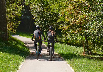 Image showing A blissful couple, adorned in professional cycling gear, enjoys a romantic bicycle ride through a park, surrounded by modern natural attractions, radiating love and happiness