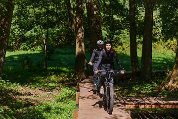 Image showing A blissful couple, adorned in professional cycling gear, enjoys a romantic bicycle ride through a park, surrounded by modern natural attractions, radiating love and happiness