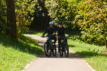 Image showing A blissful couple, adorned in professional cycling gear, enjoys a romantic bicycle ride through a park, surrounded by modern natural attractions, radiating love and happiness