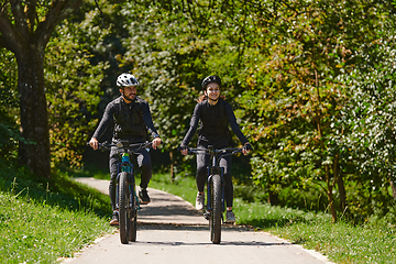 Image showing A blissful couple, adorned in professional cycling gear, enjoys a romantic bicycle ride through a park, surrounded by modern natural attractions, radiating love and happiness