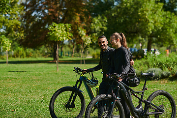 Image showing A blissful couple, adorned in professional cycling gear, enjoys a romantic bicycle ride through a park, surrounded by modern natural attractions, radiating love and happiness