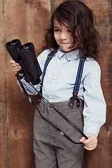 Image showing Vintage, child and boy with binoculars or fashion, style and clothes for costume on wood background or studio. Retro, kid and confidence in old fashioned spy aesthetic with shirt and suspenders