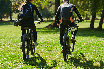 Image showing A blissful couple, adorned in professional cycling gear, enjoys a romantic bicycle ride through a park, surrounded by modern natural attractions, radiating love and happiness