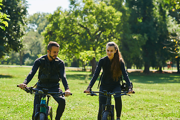 Image showing A blissful couple, adorned in professional cycling gear, enjoys a romantic bicycle ride through a park, surrounded by modern natural attractions, radiating love and happiness