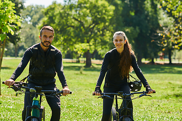 Image showing A blissful couple, adorned in professional cycling gear, enjoys a romantic bicycle ride through a park, surrounded by modern natural attractions, radiating love and happiness