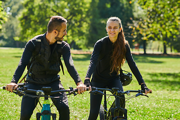 Image showing A blissful couple, adorned in professional cycling gear, enjoys a romantic bicycle ride through a park, surrounded by modern natural attractions, radiating love and happiness