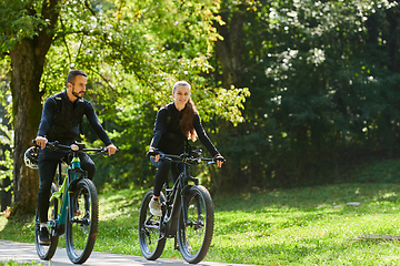 Image showing A blissful couple, adorned in professional cycling gear, enjoys a romantic bicycle ride through a park, surrounded by modern natural attractions, radiating love and happiness