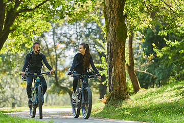 Image showing A blissful couple, adorned in professional cycling gear, enjoys a romantic bicycle ride through a park, surrounded by modern natural attractions, radiating love and happiness