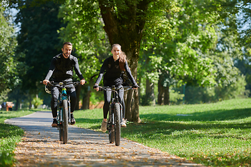 Image showing A blissful couple, adorned in professional cycling gear, enjoys a romantic bicycle ride through a park, surrounded by modern natural attractions, radiating love and happiness