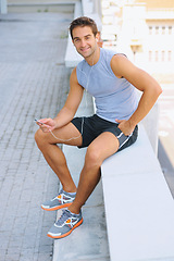 Image showing Handsome man, portrait and sitting in fitness on break from exercise, workout or outdoor training on balcony in city. Happy male person smile with earphones listening to music in rest after cardio