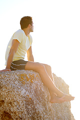 Image showing Man, sitting or rock and cliff in nature for travel, adventure or scenery with sunshine, thinking and view. Person, mountain or sky for holiday, vacation or experience outdoor and freedom or barefoot