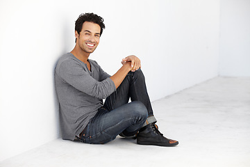 Image showing Smile, fashion and portrait of young man by a white wall in empty room with casual, cool and trendy outfit. Happy, confidence and handsome male model from Canada sitting on floor with edgy style.