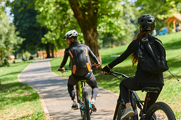 Image showing A blissful couple, adorned in professional cycling gear, enjoys a romantic bicycle ride through a park, surrounded by modern natural attractions, radiating love and happiness