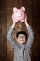 Image showing Young child, piggy bank and happy in portrait in studio, money saving and cash savings in youth with retro fashion. Boy, smile and face for coin banking and safe for bills with glasses by backdrop