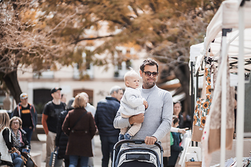 Image showing Father walking carrying his infant baby boy child and pushing stroller in crowd of people visiting Sunday flea market in Malaga, Spain.