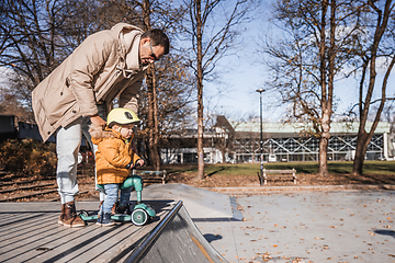 Image showing Father supervises his fearless small toddler boy while riding baby scooter outdoors in urban skate park. Child wearing yellow protective helmet