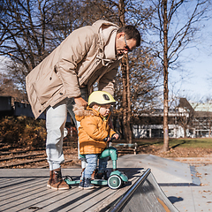 Image showing Father supervises his fearless small toddler boy while riding baby scooter outdoors in urban skate park. Child wearing yellow protective helmet