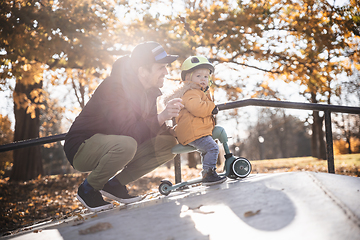 Image showing Father supervises his fearless small toddler boy while riding baby scooter outdoors in urban skate park. Child wearing yellow protective helmet