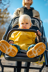 Image showing Little toddler child wearing yellow rain coat and rain boots being pushed in stroller by her mother in city park on a sunny autumn day after the rains were gone.
