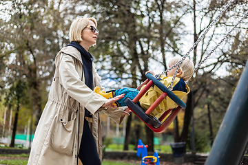 Image showing Mother pushing her infant baby boy child wearing yellow rain boots and cape on swing on playground outdoors on cold rainy overcast autumn day in Ljubljana, Slovenia