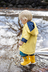 Image showing Small blond infant boy wearing yellow rubber boots and yellow waterproof raincoat walking in puddles on a overcast rainy day. Child in the rain.