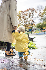 Image showing Small bond infant boy wearing yellow rubber boots and yellow waterproof raincoat walking in puddles on a overcast rainy day holding her mother's hand. Mom with small child in rain outdoors.