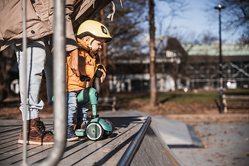 Image showing Father supervises his fearless small toddler boy while riding baby scooter outdoors in urban skate park. Child wearing yellow protective helmet