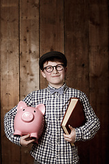 Image showing Child, piggy bank or book in thinking for education, studio background or savings for finance lesson in youth. Boy, smile and plan in coin banking of investment and learning in retro glasses fashion